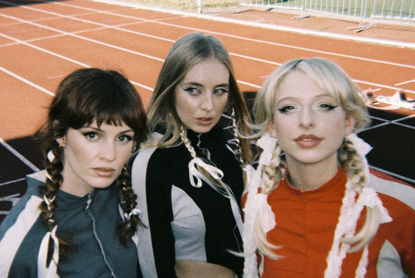Three members of the band Blusher stood on an athletics track all with plaits and bows in their hair